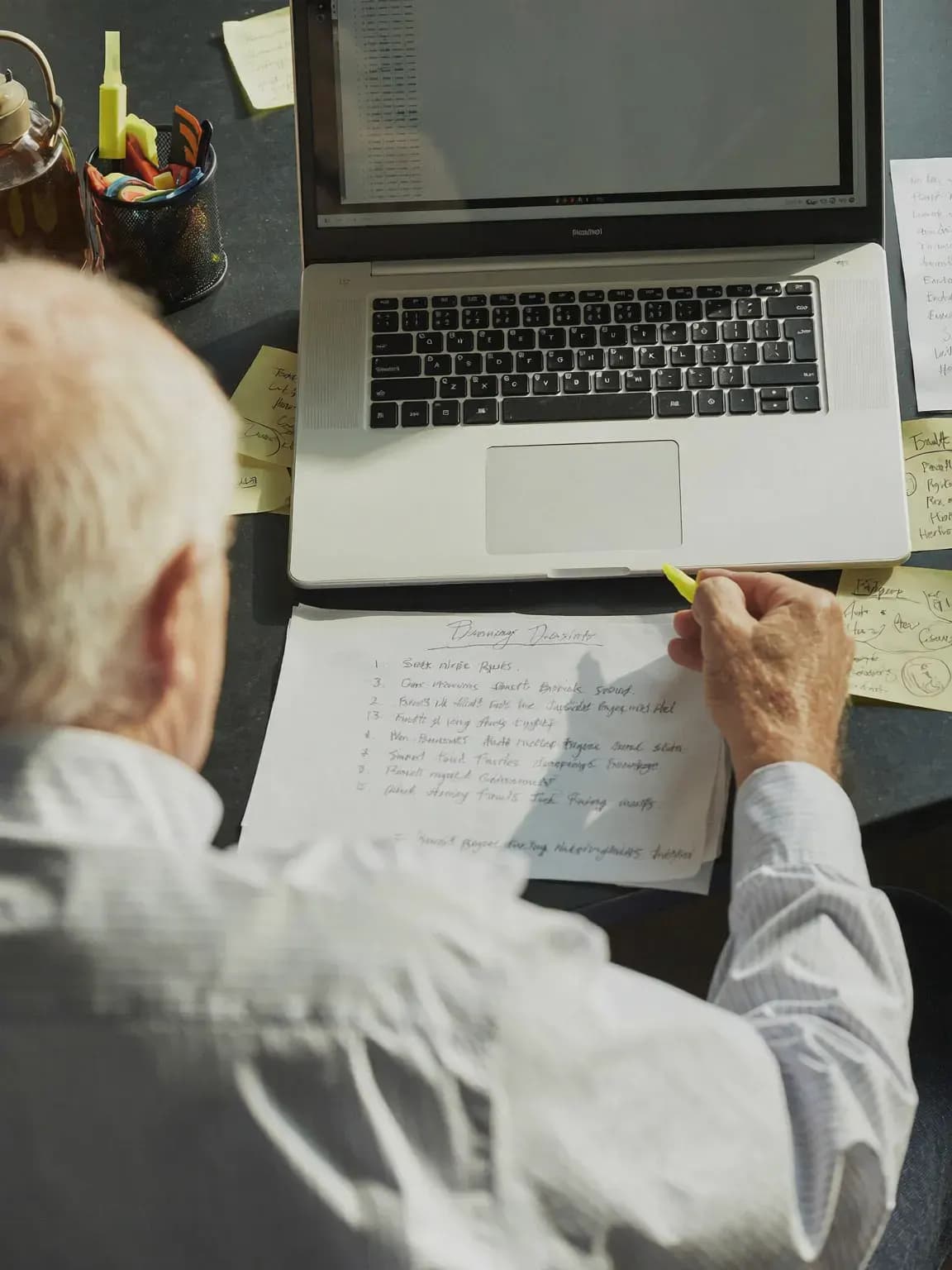 An older recruiter marking a handwritten list with a yellow highlighter next to his laptop.